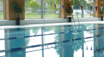 Bilden visar interiören i en simhall. En vattenspegel av en simbassäng utan badande människor i. I bagrunden är en väggyta med stora fönster.