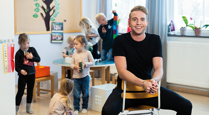 Barnskötare Robin Nord sitter i en förskolelokal med barn i bakgrunden.